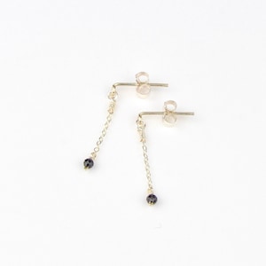 Peut inclure: Une paire de boucles d'oreilles pendantes délicates. Chaque boucle d'oreille est ornée d'une petite perle violet foncé suspendue à une fine chaîne fixée à une tige. Les boucles d'oreilles sont en métal clair, peut-être de l'or ou de l'argent, sur fond blanc.