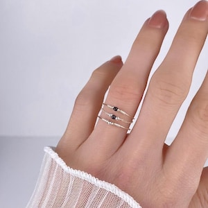 Puede incluir: Tres delicados anillos dorados, cada uno adornado con pequeñas cuentas negras, se muestran en un dedo. Los anillos son finos y de diseño minimalista, mostrando una estética simple pero elegante. El fondo es de un color suave y neutro.