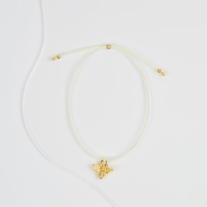 Può includere: Braccialetto delicato con un ciondolo a forma di ape dorata. Il cordino regolabile è di colore crema chiaro, con perline dorate alle estremità. Il ciondolo a forma di ape è finemente dettagliato, aggiungendo un tocco di eleganza.
