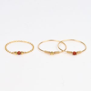 Peut inclure: Trois bagues dorées avec des accents de pierres rouge-brun. L'une est une chaîne délicate avec une pierre centrale, les deux autres sont de style créole, chacune avec une pierre et un enroulement de fil. Les bijoux sont posés sur un fond blanc.