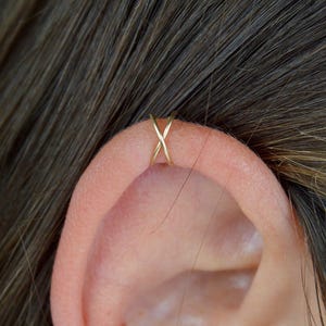 Op de afbeelding: Een gouden oormanchet in X-vorm. De manchet wordt gedragen op het bovenste kraakbeen van het oor. Het oor heeft een lichte huidskleur en het haar is donkerbruin. Eenvoudig, elegant ontwerp.