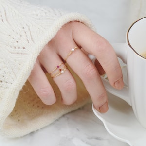 May include: A hand wearing three delicate gold rings with small gemstones. The rings feature a pearl, a red stone, and a pink stone. The hand is holding a white coffee cup.