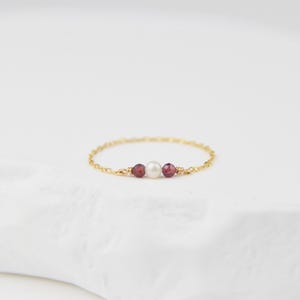 Peut inclure: Une délicate bague en chaîne dorée avec une perle blanche et deux petites pierres rouges.