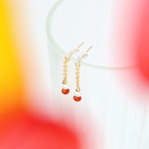 Peut inclure: Une paire de boucles d'oreilles pendantes dorées. Chaque boucle d'oreille est ornée d'une fine chaîne avec de petites perles blanches et orange. Les boucles d'oreilles sont suspendues à une tige transparente, avec un arrière-plan flou rouge, jaune et blanc.