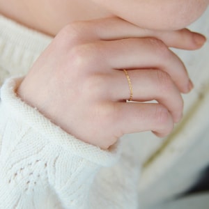 Pode incluir: Um delicado anel de corrente dourada adorna um dedo. O anel tem um design simples e elegante, adequado para uso diário. A mão repousa sobre um suéter branco de malha.