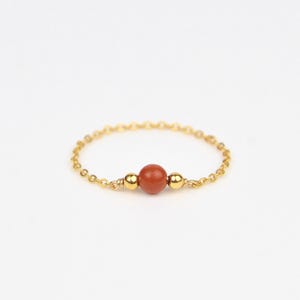 Peut inclure: Une bague dorée délicate avec une pierre précieuse centrale, ronde, brun-rouge. La monture de la bague est une fine chaîne dorée, avec une petite perle dorée de chaque côté de la pierre. Fond blanc.