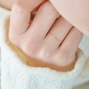 Può includere: Un delicato anello a catena d'oro indossato su un dito. L'anello è sottile e minimalista, con una lucentezza sottile. La mano è appoggiata su un maglione bianco lavorato a maglia, creando un morbido contrasto.