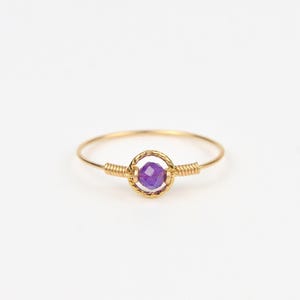 May include: A delicate gold ring featuring a vibrant purple gemstone at its center. The gemstone is encased in a circular, woven gold setting, with additional gold wire detailing on either side of the gem. The ring is set against a clean white background.