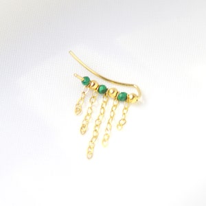 Może przedstawiać: Złoty nausznik z zakrzywionym wzorem, zielonymi koralikami i wiszącymi łańcuszkami. Nausznik ma delikatny, minimalistyczny styl, odpowiedni na różne okazje.