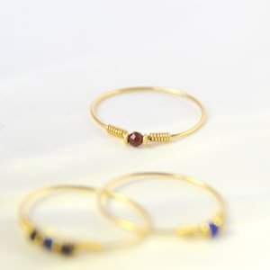Op de afbeelding: Drie gouden ringen met kleine kralen. De bovenste ring heeft een enkele donkerrode kraal, de middelste ring heeft een blauwe kraal en de onderste ring heeft een blauwe kraal.