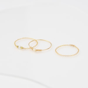 Peut inclure: Trois bagues dorées délicates. Deux bagues présentent de petites perles blanches, l'une avec un motif de chaîne et l'autre avec un détail de fil enroulé. La troisième bague est un simple motif de chaîne. Toutes les bagues sont posées sur un fond blanc.