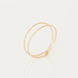 Peut inclure: Une bague dorée délicate avec un design à double bande. La bague a un style simple et minimaliste, avec deux fines bandes lisses reliées à la base. La bague est posée sur un fond blanc.