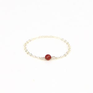 May include: A delicate silver ring with a small, faceted, red gemstone at the centre. The ring's band is a fine chain, creating a minimalist design. The gemstone is the focal point, adding a pop of colour.