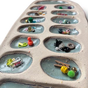 Puede incluir: Un organizador de cer&aacute;mica con m&uacute;ltiples compartimentos ovalados, cada uno con varios peque&ntilde;os suministros de artesan&iacute;a. Los art&iacute;culos incluyen botones, cuentas, pinzas para la ropa y pompones. El organizador tiene un exterior beige claro e interior azul claro.