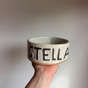 May include: A round, off-white ceramic bowl with a speckled texture and a raw, unglazed brown clay base. The word STELLA is embossed in large, bold, black capital letters around the exterior side of the bowl, creating a distinct visual contrast.