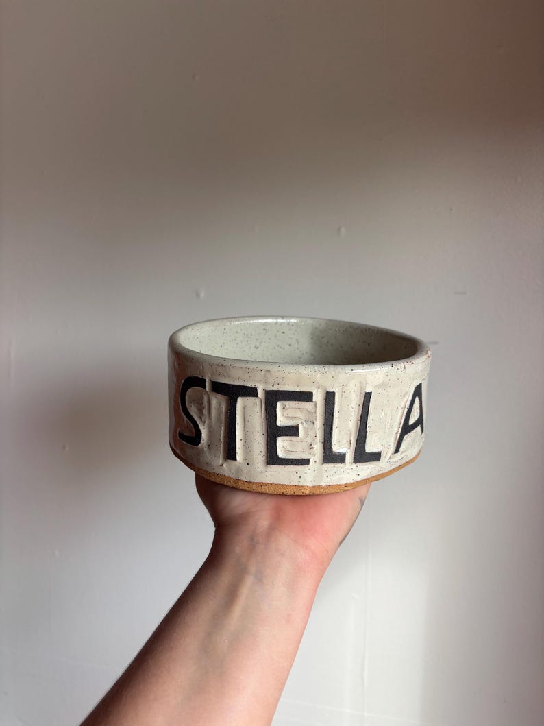May include: A round, off-white ceramic bowl with a speckled texture and a raw, unglazed brown clay base. The word STELLA is embossed in large, bold, black capital letters around the exterior side of the bowl, creating a distinct visual contrast.