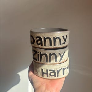 May include: A stack of three cylindrical ceramic bowls with a textured, off-white glaze. Each bowl features bold, black, debossed lettering on the exterior. The top bowl reads Danny, the middle bowl reads Zinny, and the bottom bowl reads Harry.