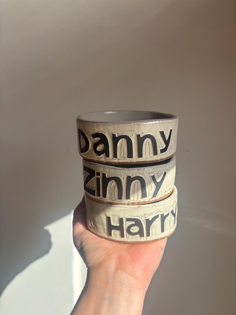 May include: A stack of three cylindrical ceramic bowls with a textured, off-white glaze. Each bowl features bold, black, debossed lettering on the exterior. The top bowl reads Danny, the middle bowl reads Zinny, and the bottom bowl reads Harry.