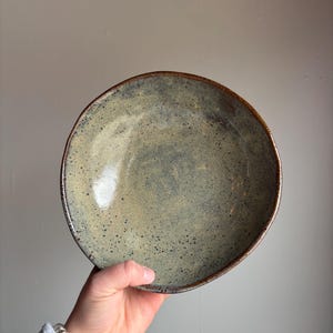 May include: A handcrafted ceramic bowl with a speckled, muted green and blue glaze. The bowl has an irregular shape and a dark brown rim. The interior has a glossy finish. The bowl is held by a person's hand.