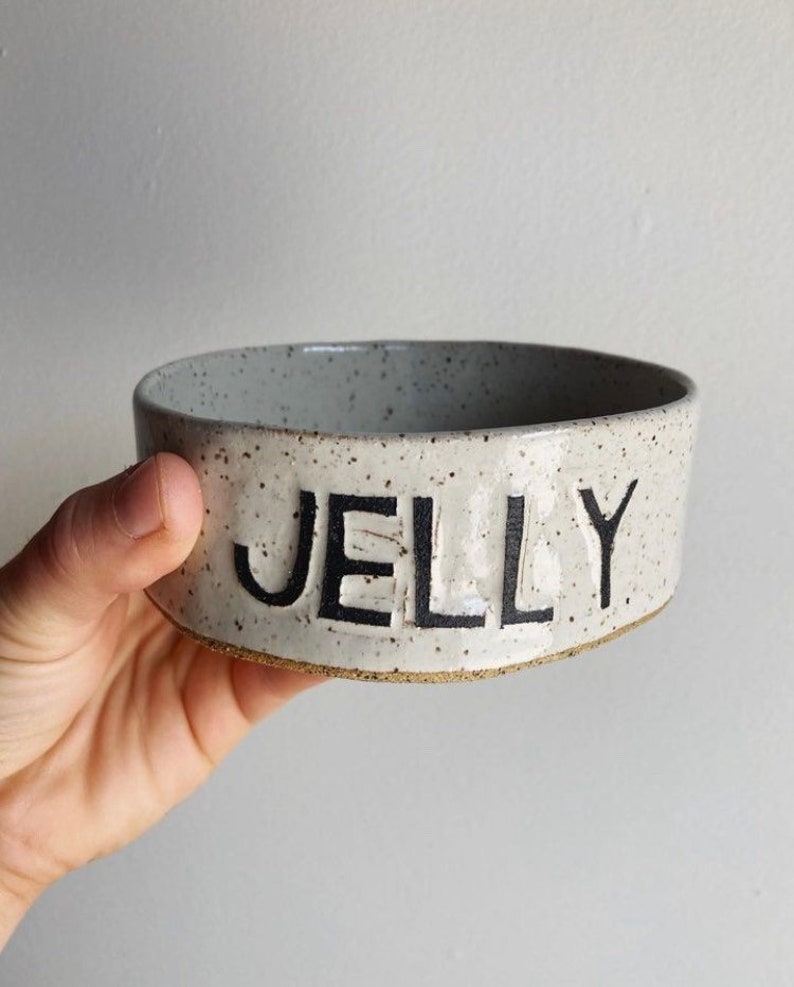 May include: A round ceramic bowl with a speckled off-white glaze and a raw clay base. The word JELLY is embossed in large, bold, black capital letters on the front exterior of the bowl. The interior of the bowl features the same speckled finish.