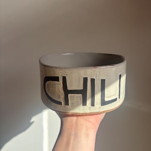 May include: A round, off-white ceramic bowl with a smooth, light-colored interior and a textured exterior. The word CHILI is embossed in large, bold, black capital letters across the front of the bowl, featuring a visible clay base rim.