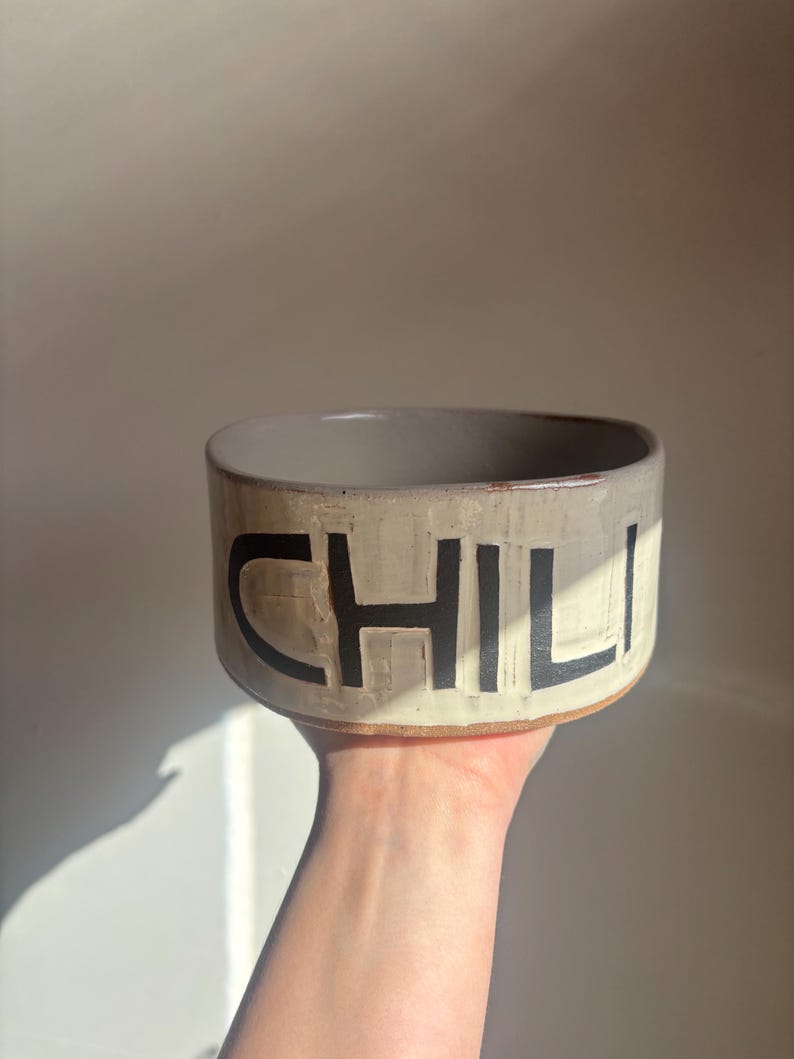 May include: A round, off-white ceramic bowl with a smooth, light-colored interior and a textured exterior. The word CHILI is embossed in large, bold, black capital letters across the front of the bowl, featuring a visible clay base rim.
