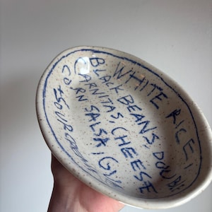 May include: Oval ceramic serving dish with a speckled, off-white surface and a blue rim. The dish features handwritten text in blue, including the words "White Rice! Double", "Black Beans, Cheese", "Carnitas, Salsa, Corn, Sour".