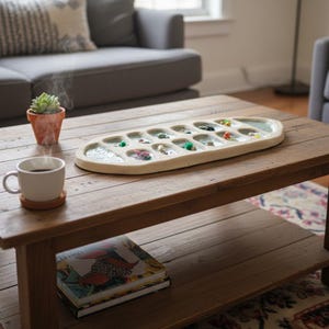 Puede incluir: Una bandeja de joyer&iacute;a ovalada de color beige claro con m&uacute;ltiples compartimentos, cada uno con varias cuentas de colores y objetos peque&ntilde;os. La bandeja est&aacute; sobre una mesa de centro de madera r&uacute;stica, junto a una taza de caf&eacute; y una peque&ntilde;a suculenta en maceta.
