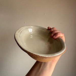 Può includere: Una ciotola in ceramica fatta a mano di forma ovale. L'interno è bianco crema, in contrasto con il bordo marrone macchiettato e strutturato. La ciotola è tenuta in mano, mostrando le sue dimensioni e il suo design.