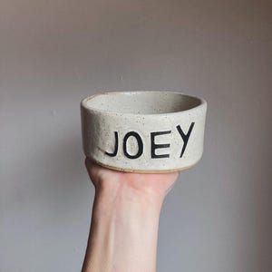 May include: A cylindrical ceramic bowl with a speckled off-white finish and a tan base. The word JOEY is embossed in large, bold, black capital letters on the front exterior of the bowl. The bowl has a thick rim and a textured, rustic appearance.
