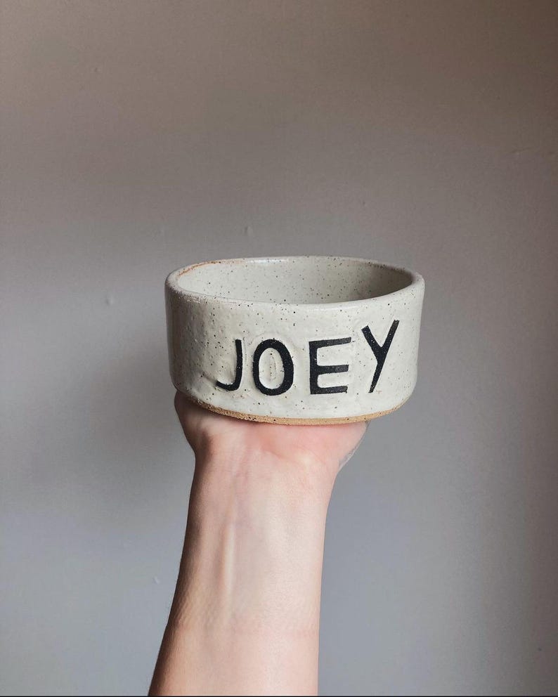 May include: A cylindrical ceramic bowl with a speckled off-white finish and a tan base. The word JOEY is embossed in large, bold, black capital letters on the front exterior of the bowl. The bowl has a thick rim and a textured, rustic appearance.