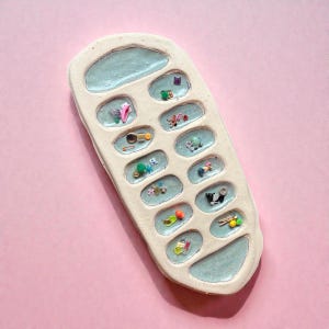 Puede incluir: Una bandeja de cer&aacute;mica en forma de tel&eacute;fono, de color crema, con detalles en azul claro. La bandeja contiene peque&ntilde;os objetos coloridos en cada uno de sus compartimentos en forma de bot&oacute;n. El fondo es rosa liso.