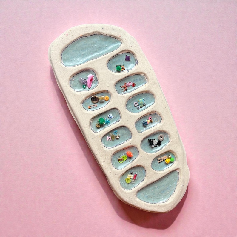 Puede incluir: Una bandeja de cer&aacute;mica en forma de tel&eacute;fono, de color crema, con detalles en azul claro. La bandeja contiene peque&ntilde;os objetos coloridos en cada uno de sus compartimentos en forma de bot&oacute;n. El fondo es rosa liso.