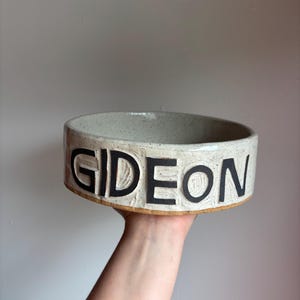 May include: A round, off-white ceramic bowl with a speckled finish and a textured surface. The word GIDEON is embossed in large, bold, black capital letters around the exterior side of the bowl. The base of the bowl shows a natural clay rim.