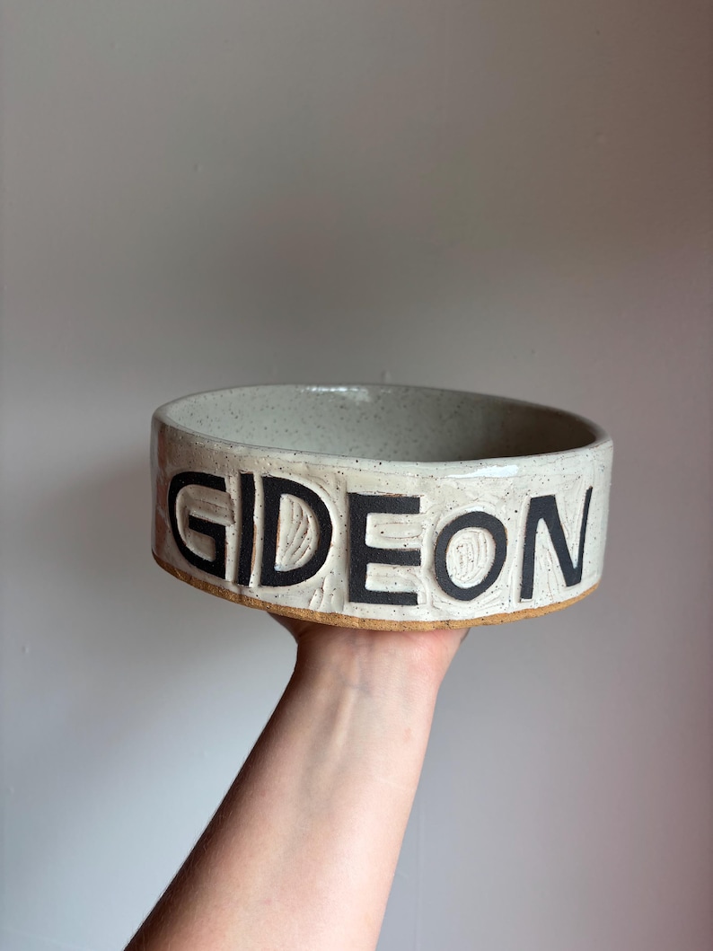 May include: A round, off-white ceramic bowl with a speckled finish and a textured surface. The word GIDEON is embossed in large, bold, black capital letters around the exterior side of the bowl. The base of the bowl shows a natural clay rim.