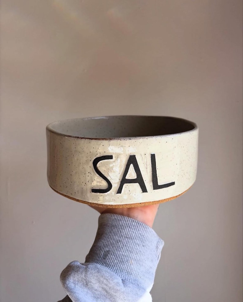 May include: A round, off-white ceramic bowl with a speckled finish and a flat base. The word SAL is printed in large, bold, black capital letters on the front exterior of the bowl. The rim is smooth and the base shows a natural, unglazed clay texture.