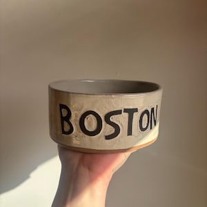 May include: A cylindrical ceramic bowl with a light beige, textured glaze and a visible terracotta-colored base. The word BOSTON is embossed in large, bold, black capital letters around the exterior side of the vessel, featuring a hand-crafted finish.