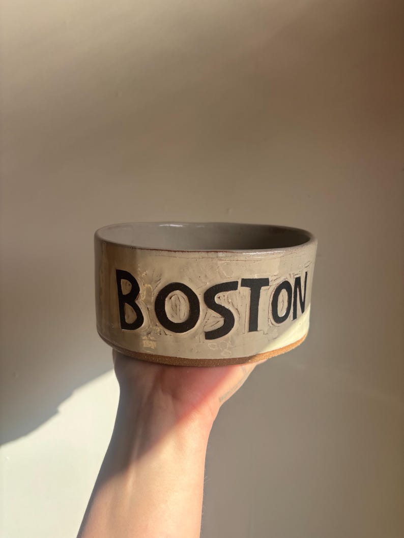 May include: A cylindrical ceramic bowl with a light beige, textured glaze and a visible terracotta-colored base. The word BOSTON is embossed in large, bold, black capital letters around the exterior side of the vessel, featuring a hand-crafted finish.