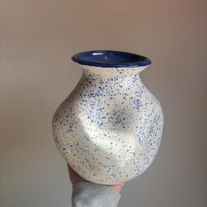 May include: A ceramic vase with a bulbous body and a flared rim. The vase is off-white with blue speckles and a dark blue interior and rim. The vase is held by a person wearing a gray sweatshirt.