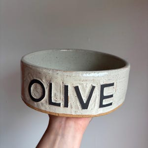 May include: A round, off-white ceramic bowl with a speckled finish and a smooth, glazed texture. The word OLIVE is embossed in large, bold, black capital letters across the front side. The base of the bowl shows the natural, unglazed brown clay.