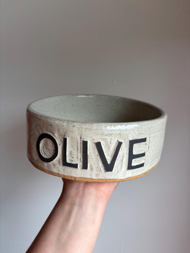 May include: A round, off-white ceramic bowl with a speckled finish and a smooth, glazed texture. The word OLIVE is embossed in large, bold, black capital letters across the front side. The base of the bowl shows the natural, unglazed brown clay.