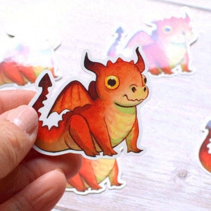 Può includere: Una collezione di adesivi di draghi colorati. Gli adesivi presentano un drago dei cartoni animati con scaglie arancioni e rosse, occhi gialli e dettagli bianchi. Un drago è tenuto in mano.