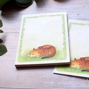 以下が含まれることがあります： 緑色の縁取りと眠っているオレンジ色のドラゴンが描かれたメモ帳用紙の2パック。メモ帳用紙の上部には「Notes」という文字が印刷されています。