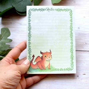以下が含まれることがあります： 緑色のノートパッドで、葉っぱの縁取りと小さなオレンジ色のドラゴンが描かれています。ノートパッドは罫線入りで、上部に「Notes」と書いてあります。