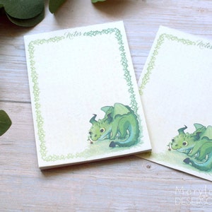 以下が含まれることがあります： 緑色のドラゴンのイラストが描かれた2つのメモ帳。メモ帳には装飾的な緑色のボーダーがあり、上部には「Notes」の文字があります。アーティストのサインが右下にあります。