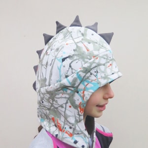 Op de afbeelding: Een witte capuchon met een kleurrijk spetterverfdesign in grijs, oranje en blauw. De hoed heeft grijze driehoekige spikes langs de bovenkant, die op een dinosaurus lijken. De hoed wordt door een persoon gedragen.