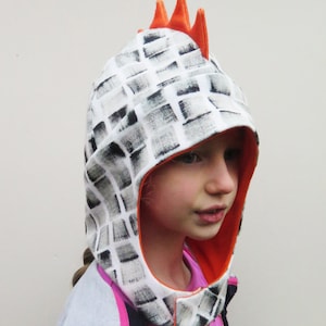 Op de afbeelding: Een kippen-thema kap voor kinderen met een wit en grijs geblokt patroon. De kap heeft een oranje binnenkant en drie oranje kammen bovenop. Het kind draagt een roze shirt en een blauw en grijs jasje.