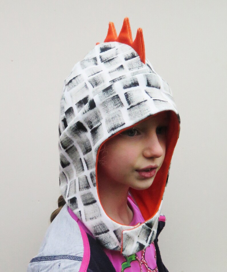 Op de afbeelding: Een kippen-thema kap voor kinderen met een wit en grijs geblokt patroon. De kap heeft een oranje binnenkant en drie oranje kammen bovenop. Het kind draagt een roze shirt en een blauw en grijs jasje.