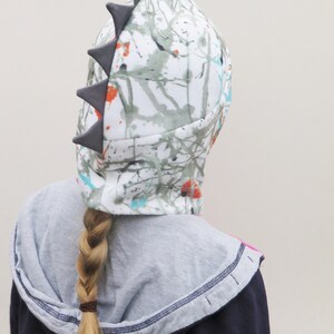 Op de afbeelding: Een witte hoed met een kleurrijk spetterverfdesign en grijze driehoekige spikes. De hoed wordt gedragen over een grijze hoodie en een donkerblauwe jas. Een blonde vlecht is zichtbaar.