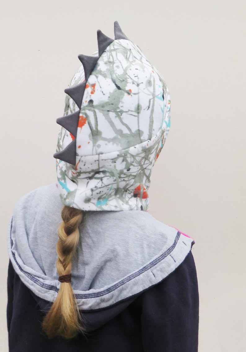 Op de afbeelding: Een witte hoed met een kleurrijk spetterverfdesign en grijze driehoekige spikes. De hoed wordt gedragen over een grijze hoodie en een donkerblauwe jas. Een blonde vlecht is zichtbaar.